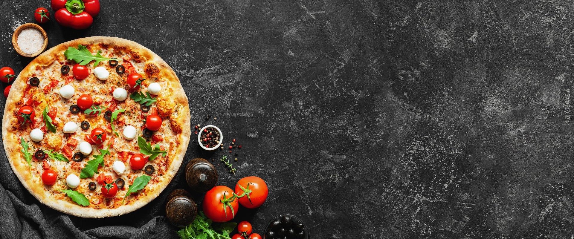 Italian pizza and pizza cooking ingredients on black concrete background. Tomatoes on vine, mozzarella, black olives, herbs and spices. Copy space for text. Banner composition